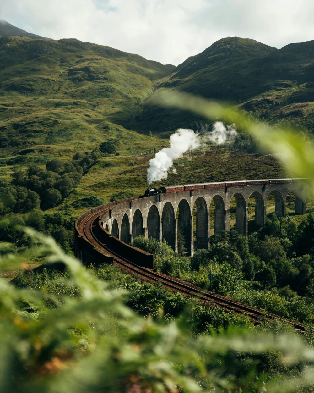 groen schots berglandschap met viaduct met daarop een trein met stoomlocomotief en rode wagons net als de hogwarts express